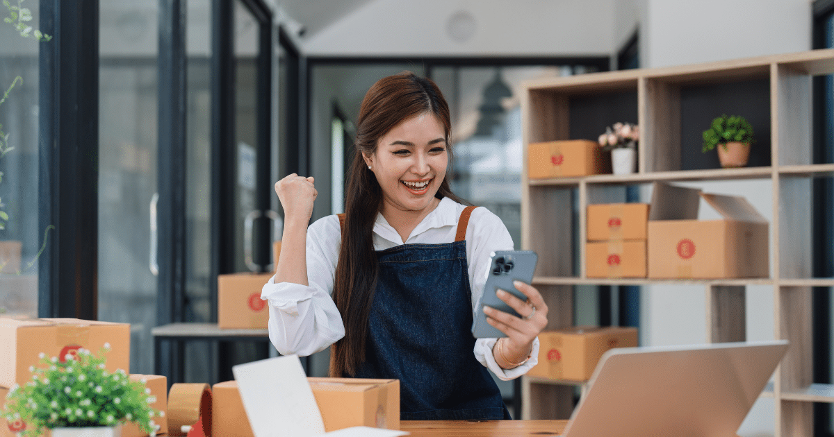 A cheerful small business owner wearing an apron celebrates a success while holding a smartphone, surrounded by neatly packed boxes and a laptop on a desk.