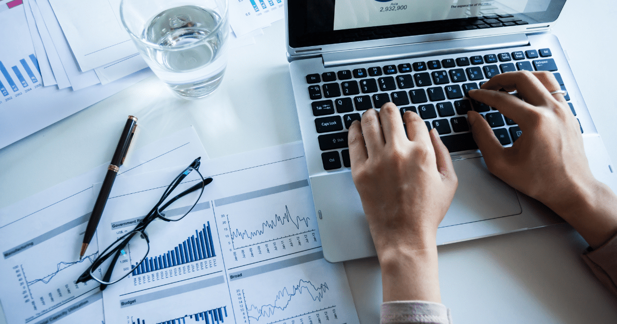 Hands typing on a laptop keyboard with financial documents, charts, and a pen on the desk.